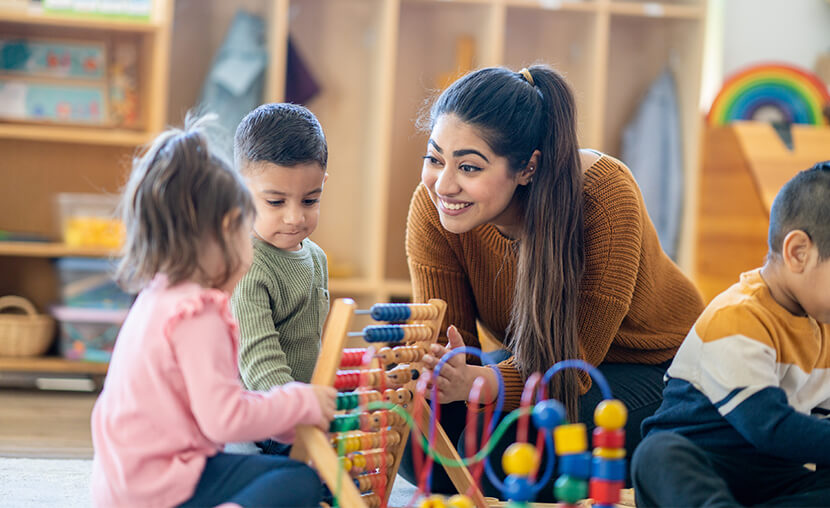 A female Kindergarten teacher sits on the floor with students as they play with various toys and engage in different activities.